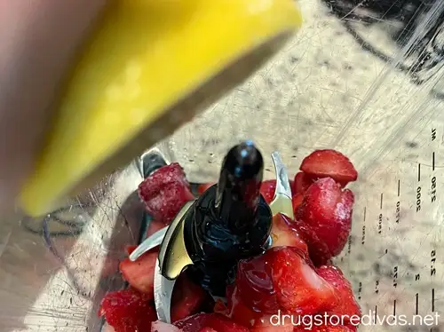 A hand squeezing half a lemon into a blender that has strawberries in it.