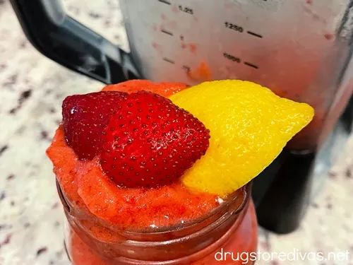 A glass of frozen strawberry lemonade with strawberry and lemon pieces on top next to a blender.