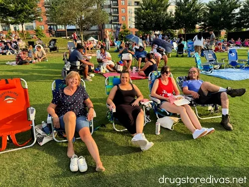 Four people, three women and a man, sitting on collapsible chairs while surrounded by other people on a lawn at Live Oak Bank Pavilion.