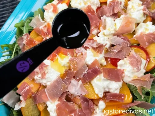 A measuring spoon filled with liquid over sliced prosciutto, burrata filling, and peach slices over arugula in a blue tray.