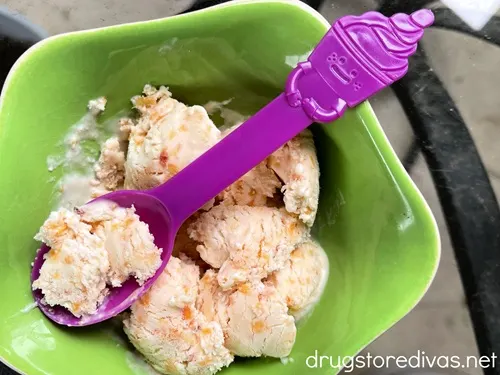 Peach ice cream scoops in a green bowl with a blue spoon that has a cartoon ice cream cup on top.