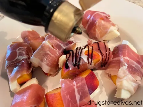 Balsamic glaze in a bottle being drizzled on top of a tray of prosciutto wrapped peach and mozzarella slices.