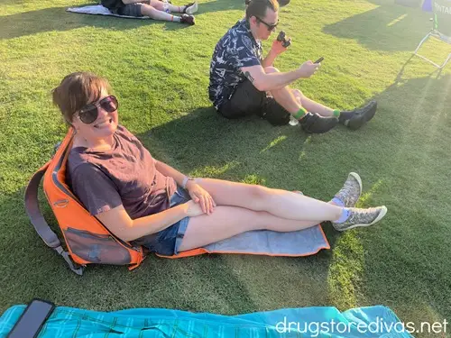 A woman in a t-shirt and shorts sitting on a blanket with a chair-like back on the ground at an outdoor music venue with a man sitting and holding a fan towards himself behind her.