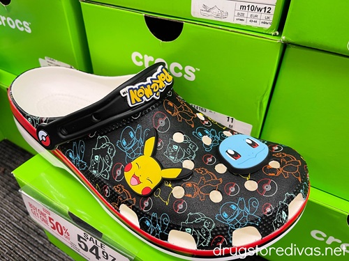 A pair of black Croc shoes with a Pikachu and Squirtle jibbitz on it with green Croc boxes in the background.