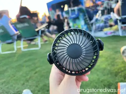 A hand holding a portable fan while sitting in lawn seats at an outdoor concert venue.