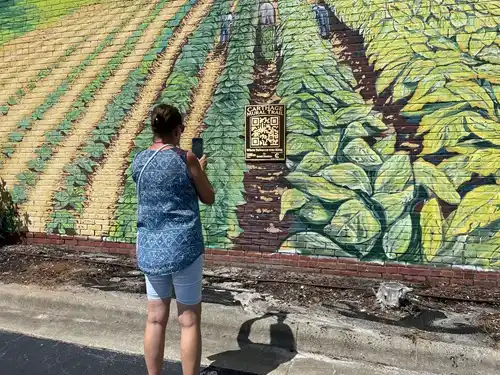 A woman looking at a mural with her phone up scanning a metal QR code on the mural.