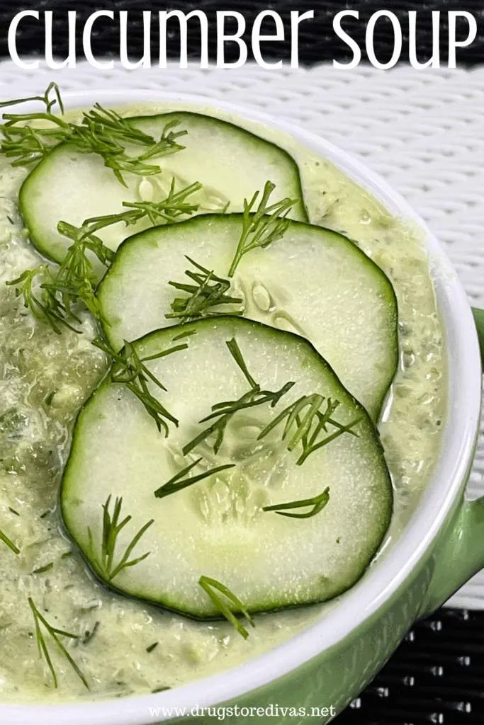 A green bowl with a white rim filled with a green soup with cucumber slices and dill on top and the words "Cucumber Soup" digitally written on top.