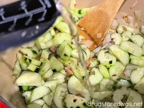 Liquid being pored from a measuring cup onto chopped onion, garlic, and sliced cucumbers in a Dutch oven with a spoon.