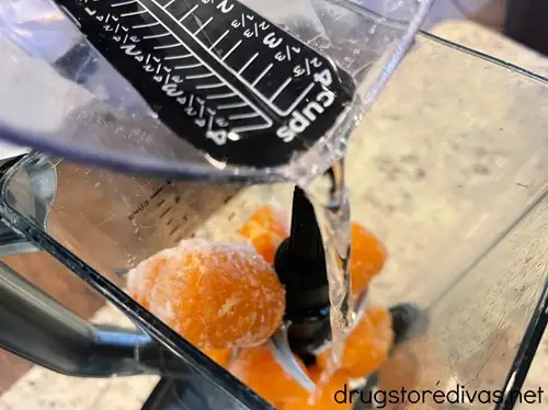 Water in a measuring cup being poured over mandarin oranges in a blender.