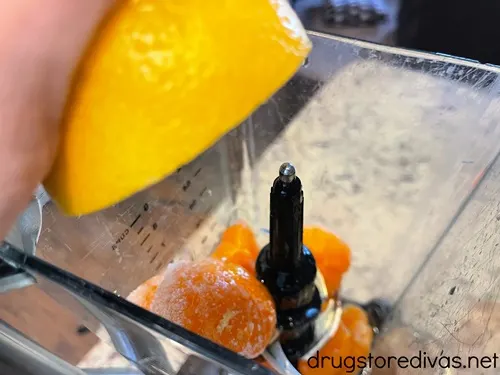 A hand squeezing half a lemon into a blender that's filled with mandarin oranges.