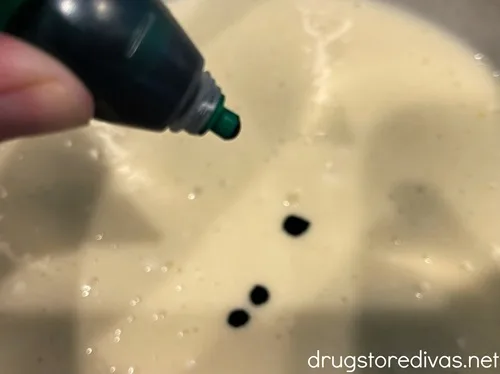A hand adding green food coloring to a white batter with three green food coloring dots in it.