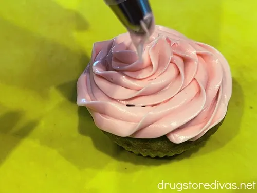 Pink icing being piped on top of a green cupcake that's resting on a green cutting board.