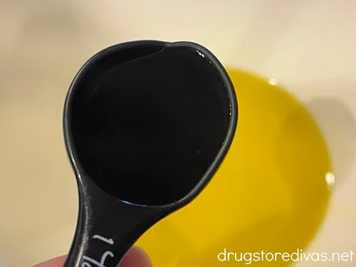 A measuring spoon above a white bowl that has olive oil in it.