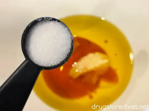 A measuring spoon with salt above a white bowl that has olive oil in it.