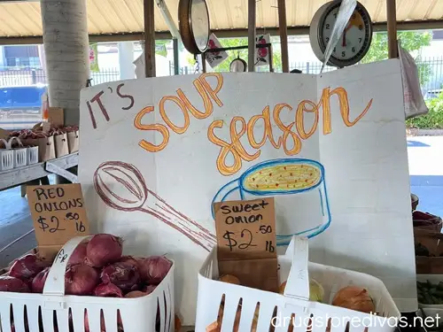 A large sign that says "It's Soup Season" in front of onions for sale at the Nashville Farmers Market.