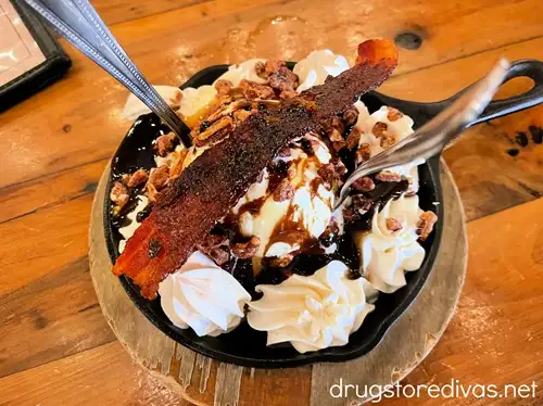 A fried brownie dessert with whipped cream and a candied piece of bacon on top, plus two spoons.