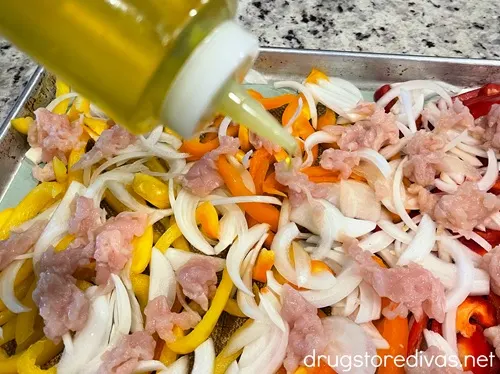 A squeeze bottle of oil above a sheet pan filled with pieces of raw chicken, sliced onion, and yellow, orange, and red peppers.