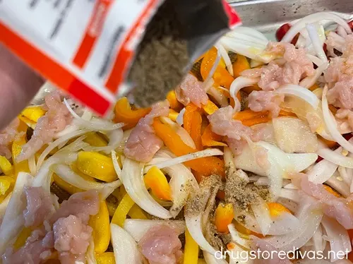A packet of seasoning above a sheet pan filled with pieces of raw chicken, sliced onion, and yellow, orange, and red peppers.