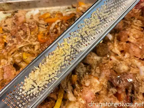 Garlic on a microplane grater above a sheet pan filled with pieces of raw chicken, sliced onion, and yellow, orange, and red peppers.