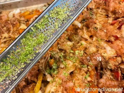 Lime zest on a microplane grater above a sheet pan filled with pieces of raw chicken, sliced onion, and yellow, orange, and red peppers.