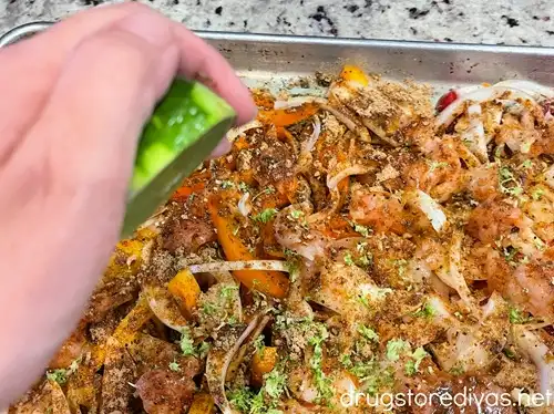 A hand squeezing half a lime above a sheet pan filled with pieces of raw chicken, sliced onion, and yellow, orange, and red peppers.