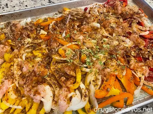 Pieces of raw chicken, sliced onion, and yellow, orange, and red peppers with seasoning on top on a sheet pan.