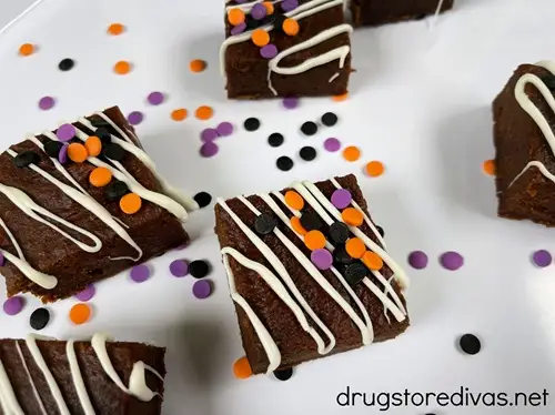 Brownies decorated with white lines and black, orange, and purple circle sprinkles, with those same sprinkles around them, on a white tray.