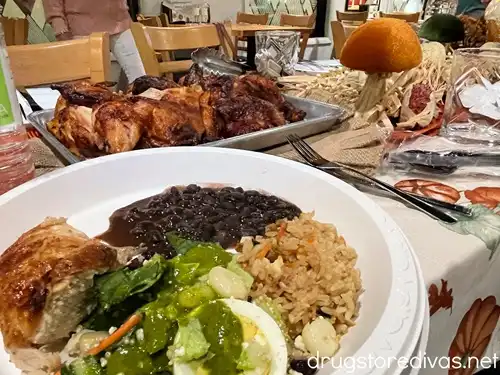 A plate with chicken, black beans, fried rice, and salad on it with a tray of chicken behind it.