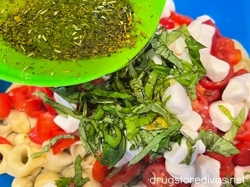 Dressing in a green bowl being poured over sliced basil, mozzarella pearls, and sliced cherry tomatoes and cooked tortellini in a blue bowl.