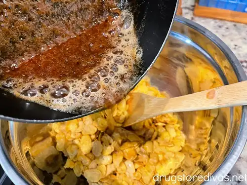A toffee mixture bring poured over corn flakes in a silver bowl with a bamboo spoon also in the bowl.