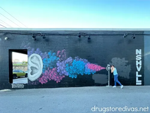 A woman in a blue tank top and blue jeans posting in front of a colorful mural painted on a wall.