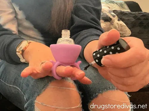 A woman wearing a purple nail polish holder on one hand and holding the top of a nail polish bottle in the other.