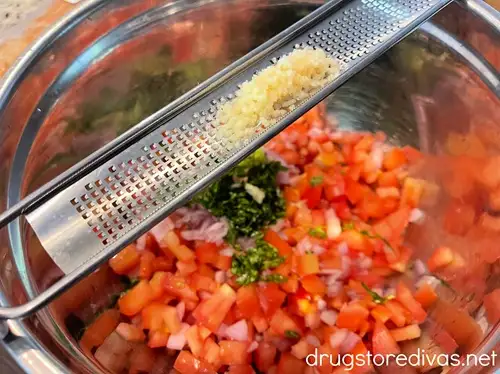 Fresh garlic on a microplane grater above a silver bowl with diced tomatoes, shallots, and chopped basil.
