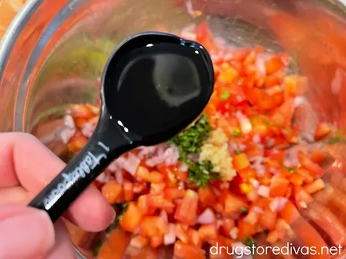 A measuring spoon with liquid above a silver bowl with diced tomatoes, shallots, and chopped basil.