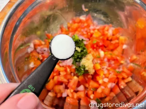 A measuring spoon with salt above a silver bowl with diced tomatoes, shallots, and chopped basil.