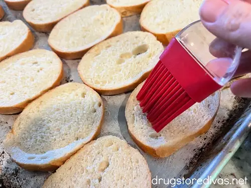 A red pastry brush brushing olive oil onto sliced bread on a baking sheet.