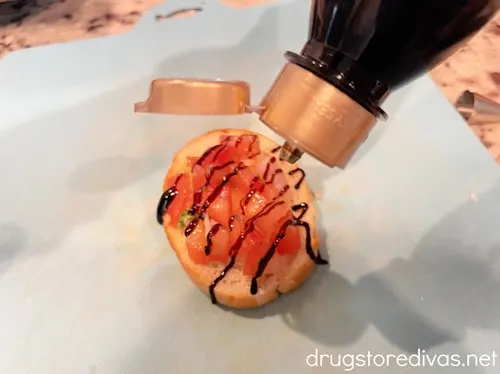 A bottle of balasamic glaze piping lines on top of a bruschetta topped piece of bread.