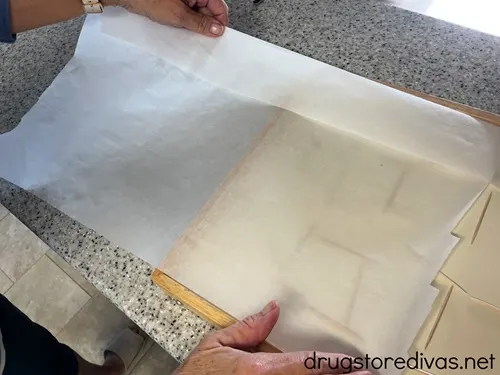 Hands laying parchment paper on top of a rectangular board on a counter.