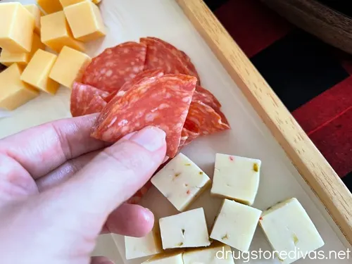 A piece of salami being folded and held by a hand above a board, which has two piles of cubed cheese on it.