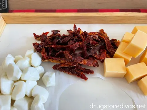 Mozzarella pearls, sun-dried tomatoes, and an orange cheese on a board.