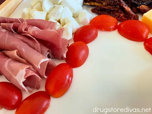 Cherry tomato halves making a boarder around prosciutto and mozzarella pearls that are on a board.