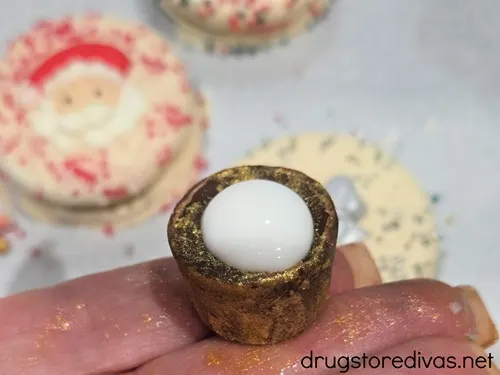 A Rolo chocolate with white chocolate on it balancing on fingers with cookies in the background.