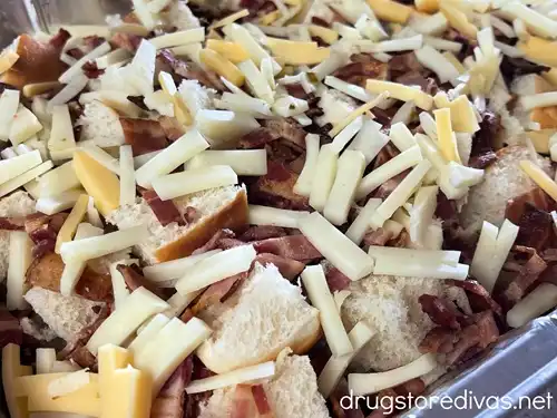 Pieces of cheese and bacon on top of bread cubes in an aluminum pan.