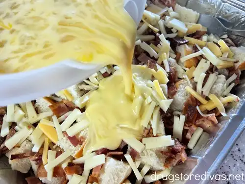 An egg mixture being poured over bread cubes, chopped cheese, and chopped bacon in an aluminum tray.