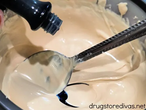 Blue food coloring being poured into a bowl with melted white chocolate and a spoon.