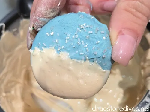 A hand holding a chocolate dipped Oreo cookie that's light blue with sugar sprinkles on top and white at the bottom.