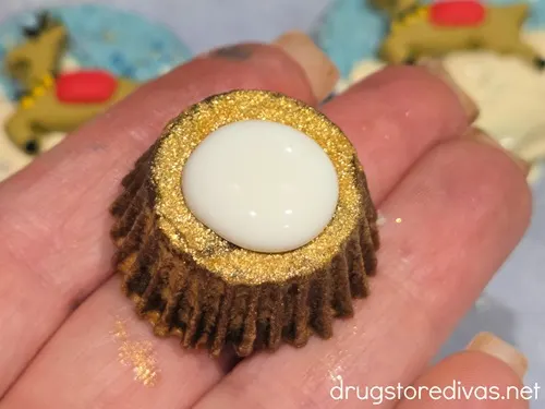 A hand holding a gold glittered mini Reese's peanut butter cup with a dab of white chocolate in the middle.