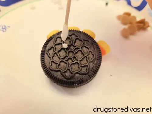 A toothpick with white chocolate on it above an Oreo cookie that has four pieces of candy corn sticking out of it.