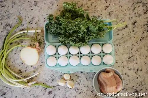 Green onions, half a white onion, kale, a dozen eggs, garlic cloves, and bacon in a bowl on a table.