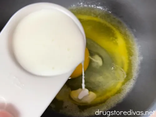 A white measuring spoon pouring cream into a gray bowl with a raw egg at the bottom.